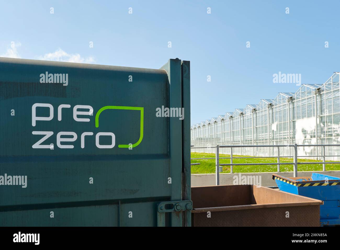 Contenitore PreZero vicino a un'area di serra industriale. PreZero è un'azienda di trattamento dei rifiuti che pone l'accento sulla sostenibilità e il riciclaggio Foto Stock