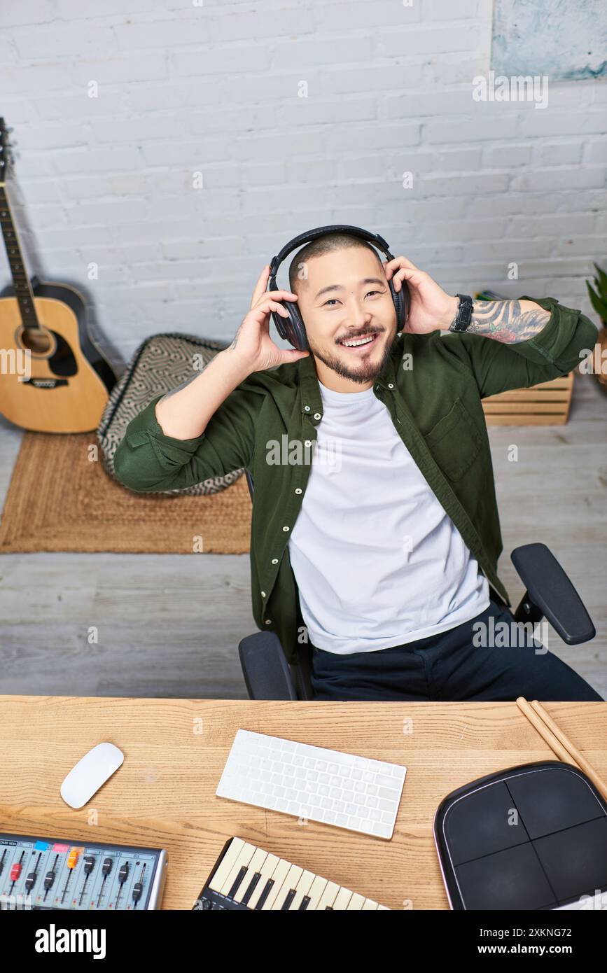 Un giovane asiatico con una camicia verde sorride mentre ascolta la musica attraverso le sue cuffie nel suo studio di registrazione domestico. Foto Stock