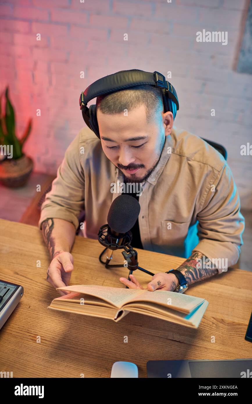 Un giovane asiatico siede in uno studio di podcast, indossa le cuffie, tenendo un libro aperto mentre registra un episodio. Foto Stock