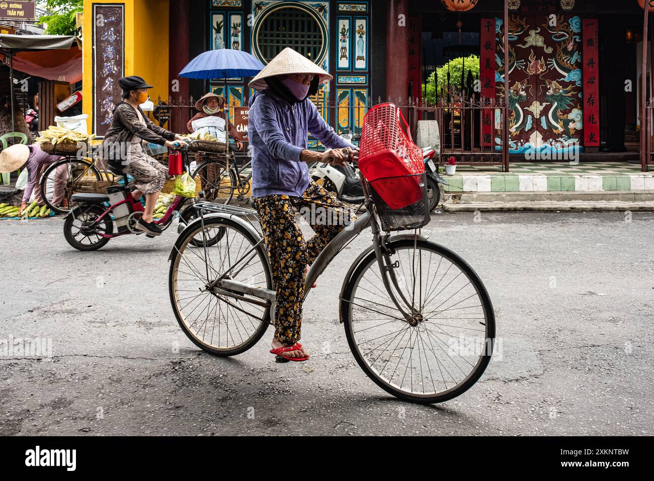 Una donna anziana con un cappello conico in bicicletta a Hoi An Vietnam. Donna vietnamita in tradizionale cappello di bambù in bicicletta lungo l'antica città di Hoi An. Trav Foto Stock