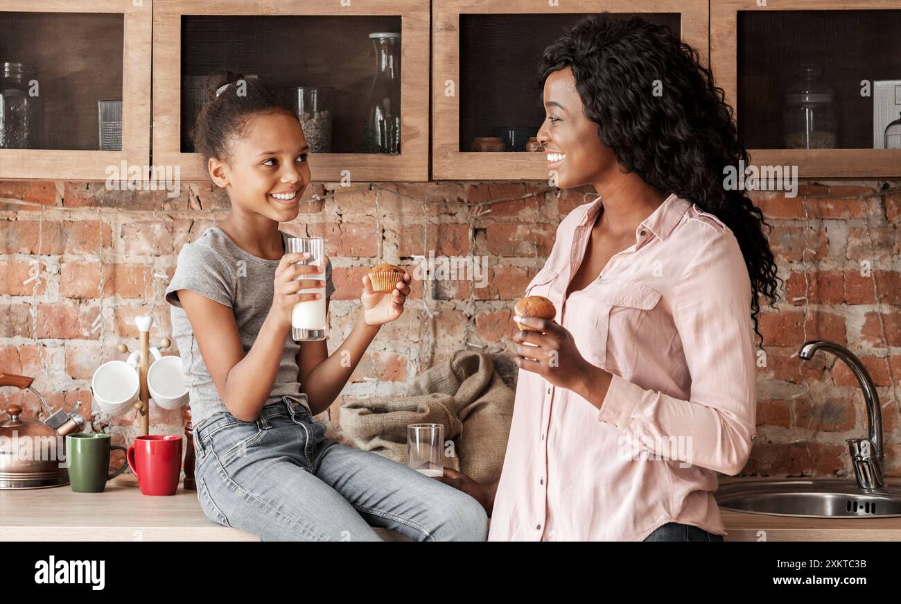 Allegra madre e figlia africana che passano del tempo insieme, bevono latte e mangiano cupcake in cucina Foto Stock