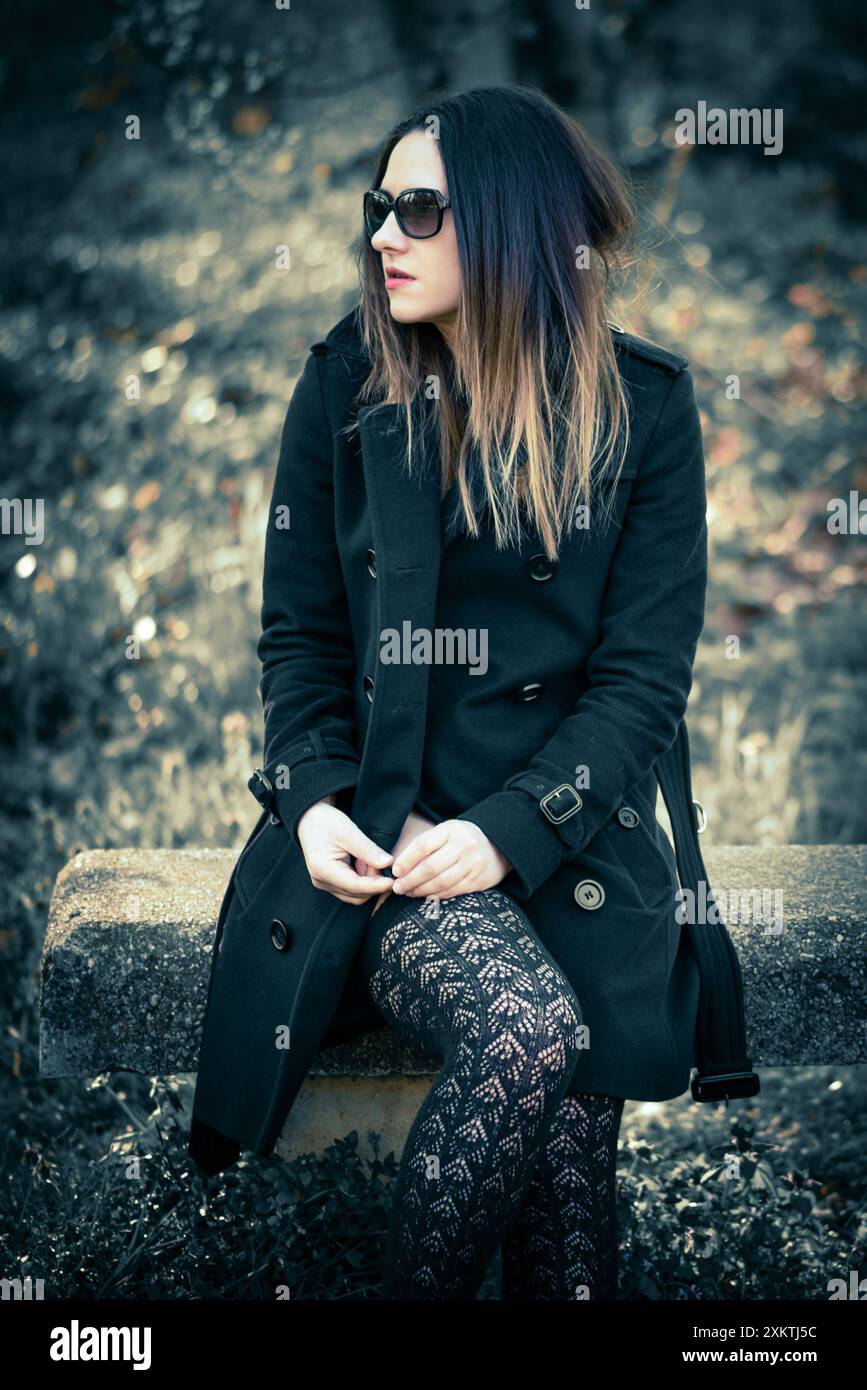Una giovane donna che indossa un cappotto nero e calze alte sulla coscia si siede su un tronco intempestivo in un ambiente forestale. I suoi capelli scuri le incorniciano il viso e lei indossa Foto Stock
