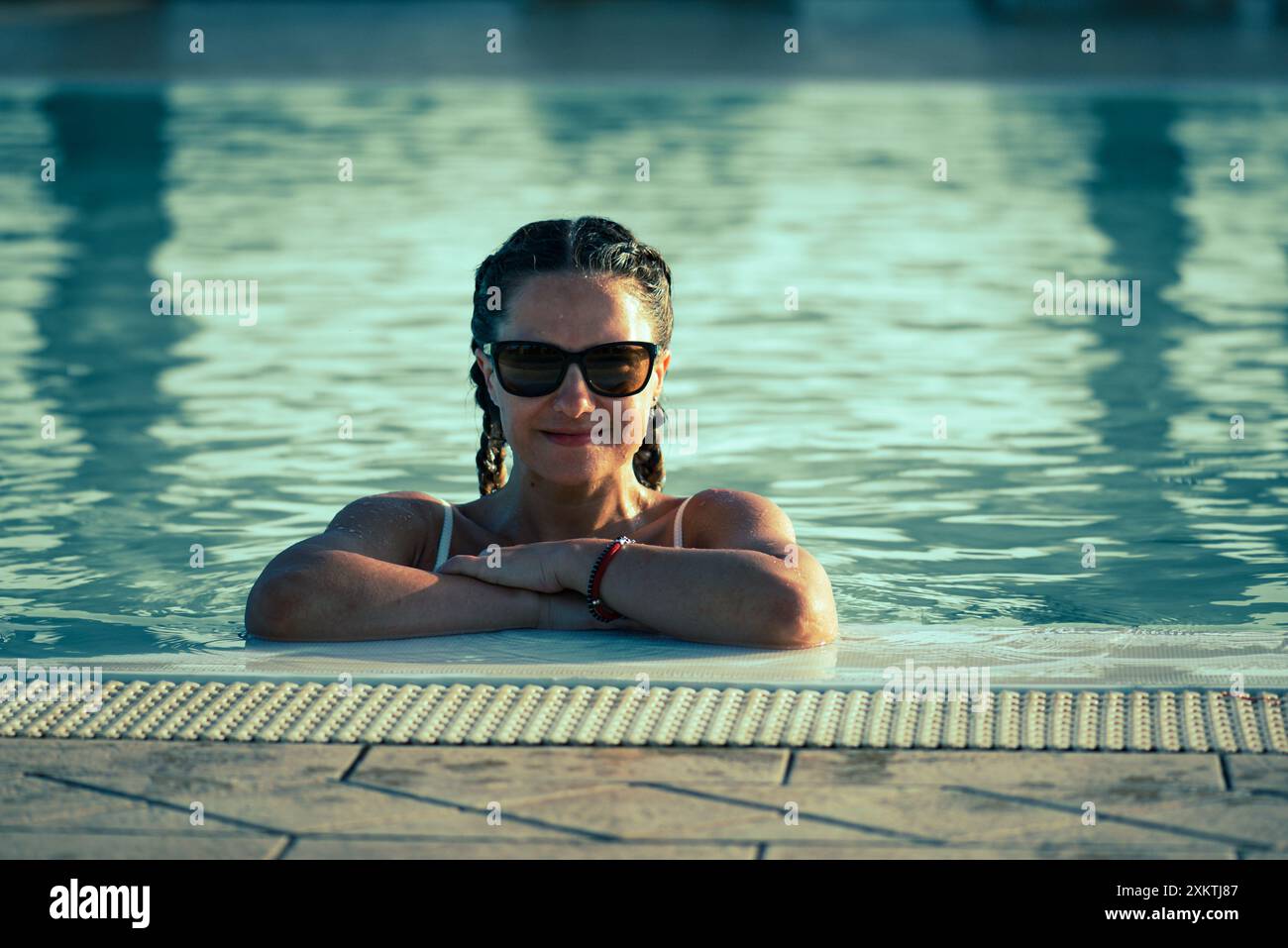 Una donna che indossa gli occhiali da sole poggia le braccia sul bordo di una piscina, con il viso e la parte superiore del corpo sopra l'acqua. Sembra che sia rilassata e goda Foto Stock