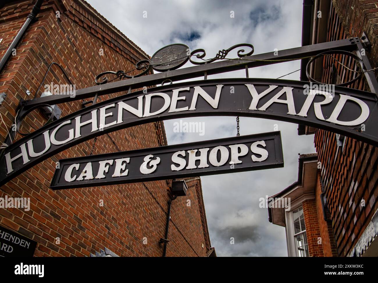 Un cartello ad arco con lo 'Hughenden Yard Cafe & Shops' adagiato su un cielo parzialmente nuvoloso, con edifici in mattoni su entrambi i lati. Foto Stock