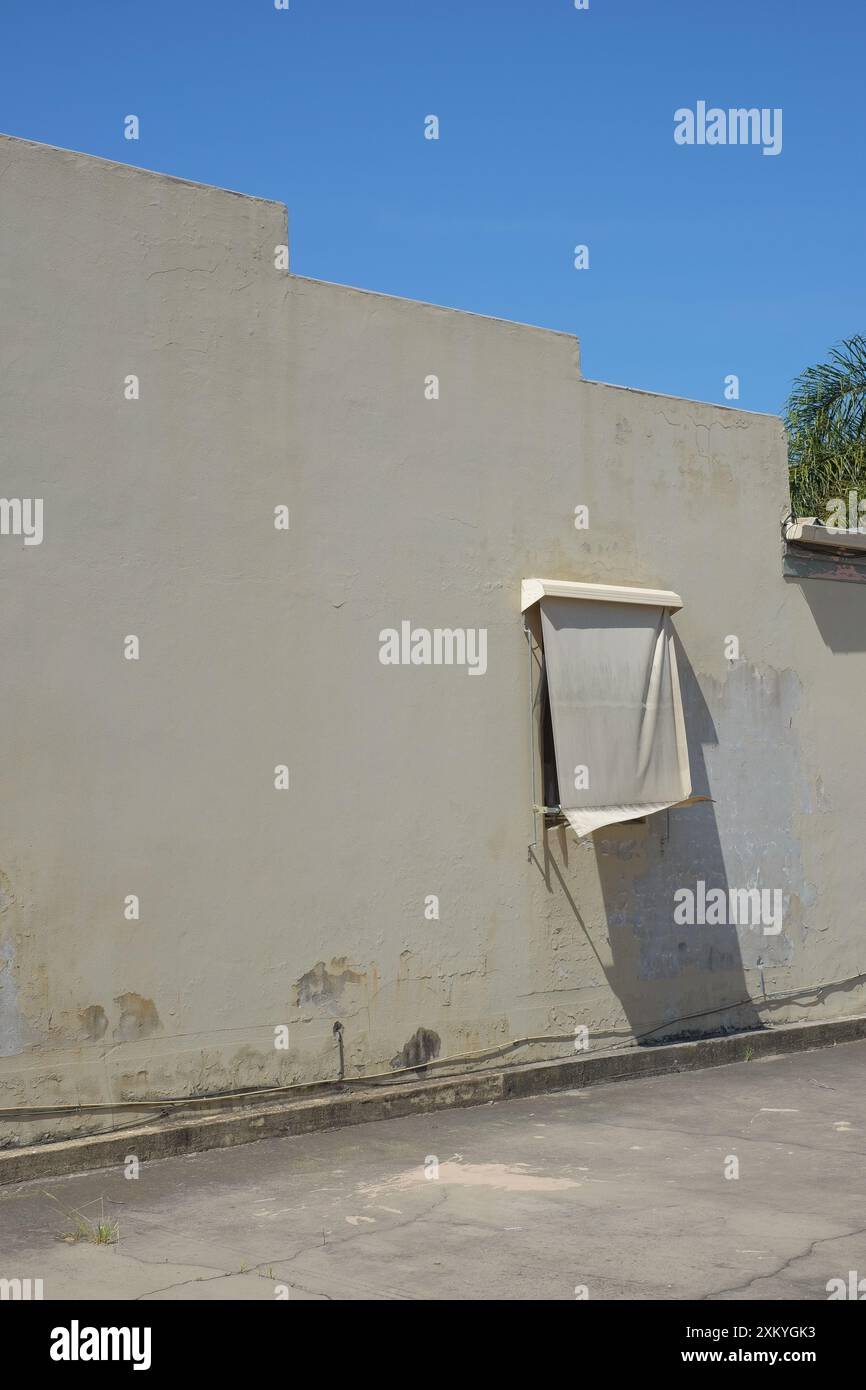 Una finestra tagliata in un muro coperto da un panno beige esterno cieco dello stesso colore della parete, un parapetto discendente a gradini contro il cielo blu Foto Stock