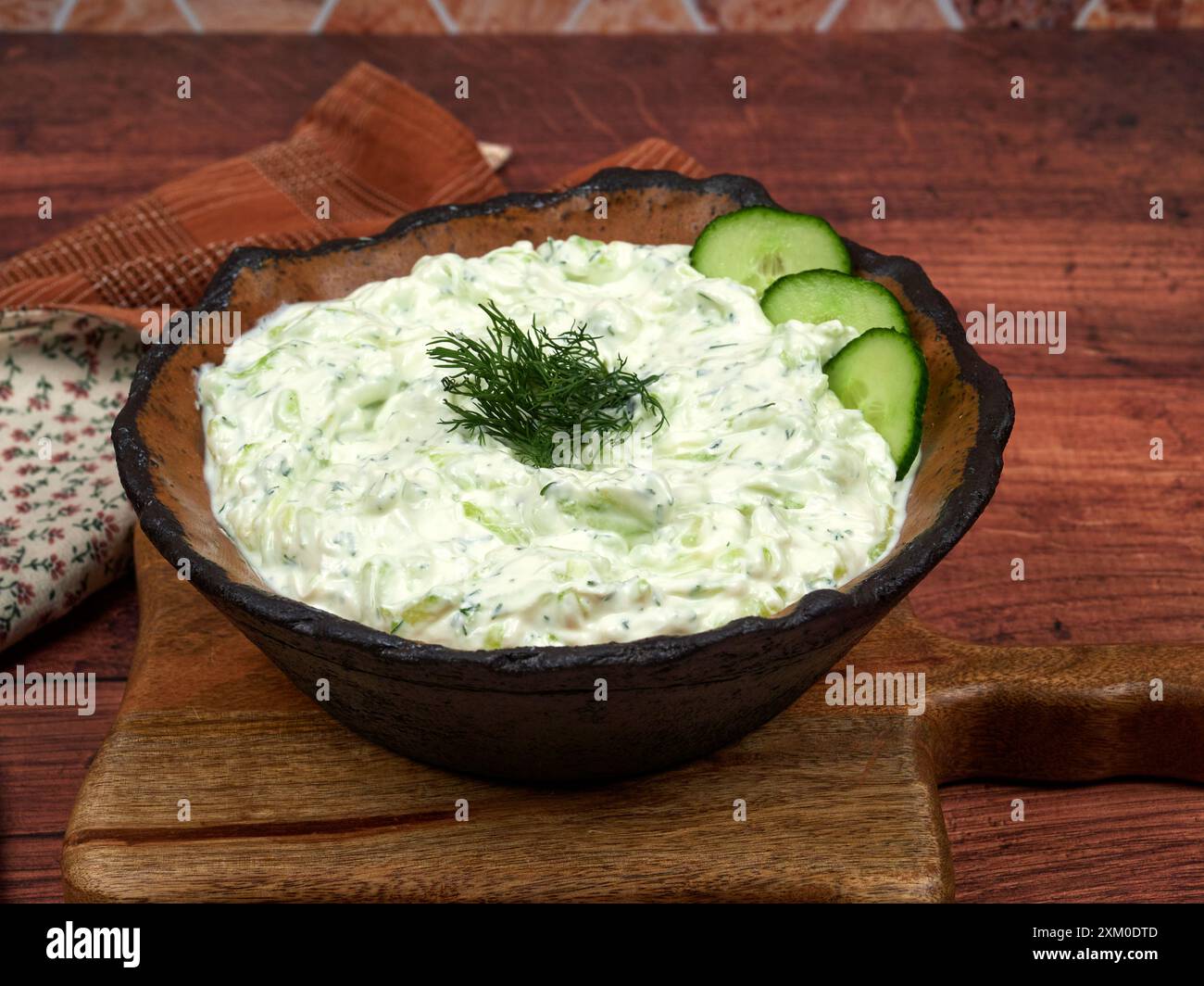 Una ciotola di Tzatziki, una cremosa salsa di yogurt greco con cetrioli tagliati finemente, aglio e aneto, adagiata su un tagliere di legno, con un panno a motivi geometrici Foto Stock