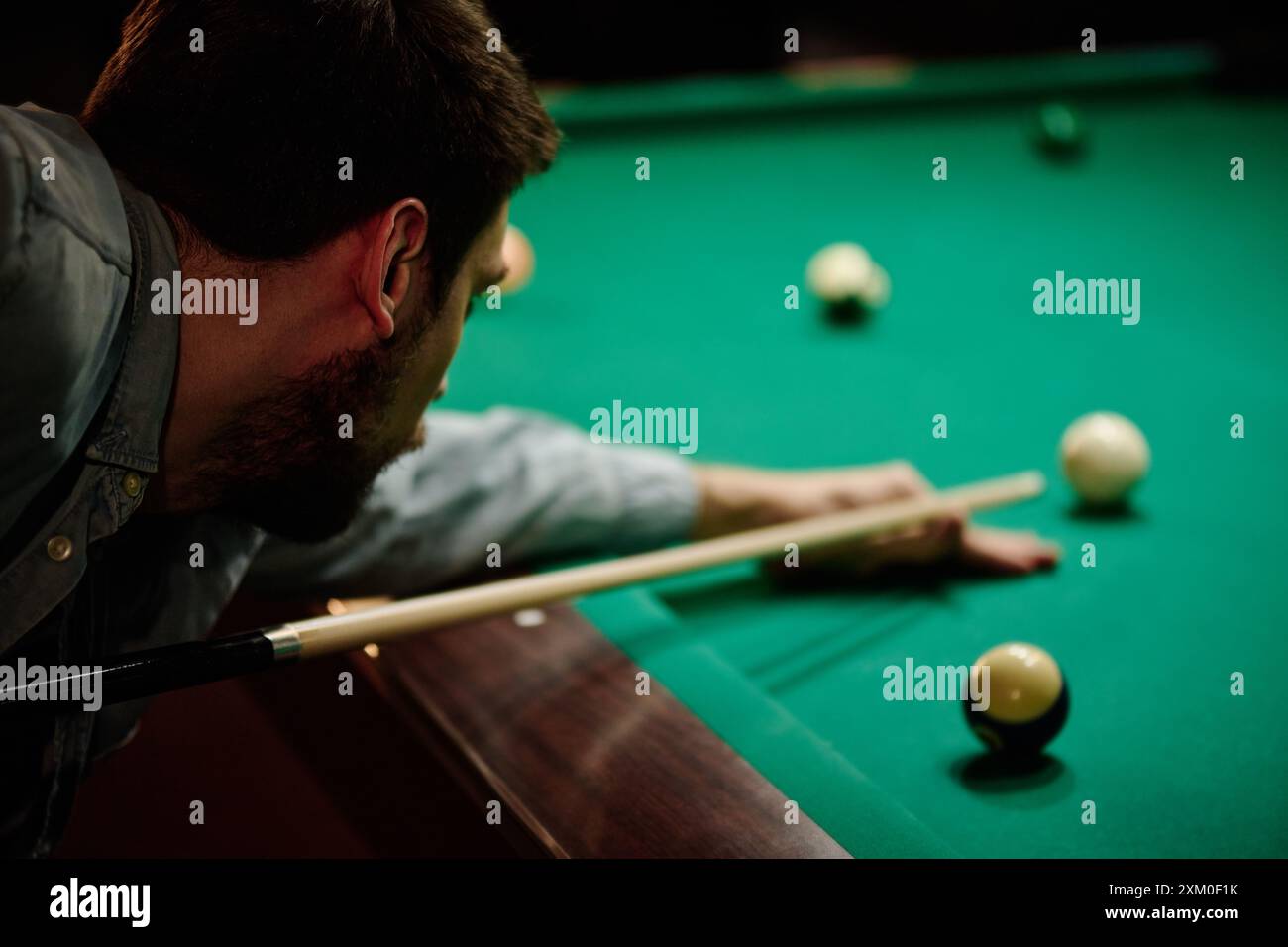 Concentrati su un giovane giocatore di biliardo maschile in abbigliamento casual che tiene in mano il segno mentre si piega sul tavolo verde e gioca nella sala giochi Foto Stock