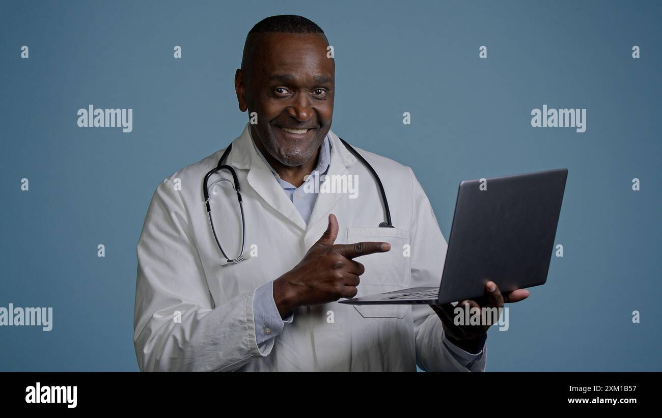 Uomo afroamericano medico terapeuta chirurgo maschile etnico in cappotto bianco in studio sfondo blu che lavora su laptop consulenza paziente on-line remoto Foto Stock