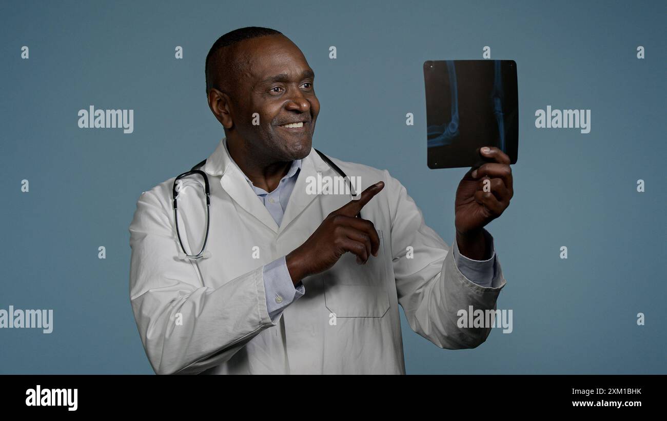 Pensivo sorridente uomo afroamericano maschio radiologo medico chirurgo traumatologo che esamina radiografia in studio blu background check risultati scansione Foto Stock