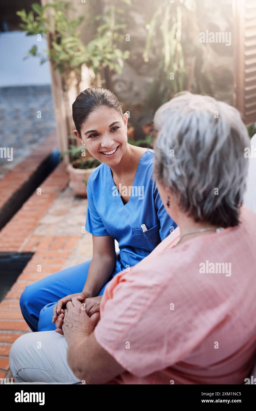 Tenendosi per mano, felice e badante con una donna anziana in giardino per terapia, gentilezza e chiacchierare. Salute mentale, discussione e operatore medico o supporto Foto Stock
