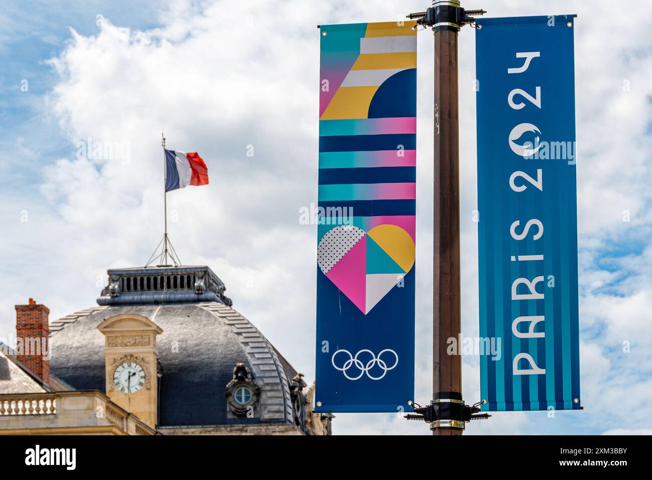 Cartelli decorativi colorati per le Olimpiadi estive di Parigi 2024 in una strada di Parigi, in Francia, con una bandiera francese che sventola su un tetto sullo sfondo Foto Stock