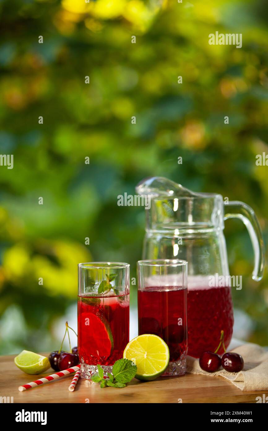 Una caraffa di bicchiere di punch rinfrescante alla frutta, accompagnata da due bicchieri guarniti con lime, menta e ciliegie. Foto verticale. Foto Stock