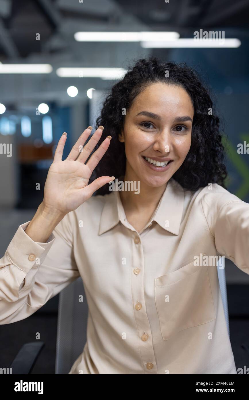 Donna sorridente che salta durante le videochiamate in un ufficio moderno. Un gesto amichevole crea interazioni accoglienti. Ambiente professionale con comunicazione multimediale che favorisce le connessioni sul luogo di lavoro. Foto Stock