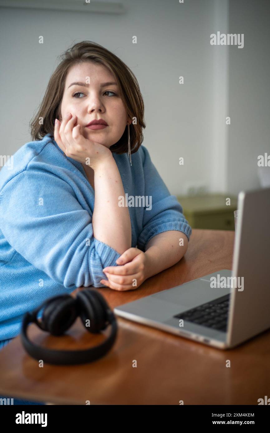 La giovane donna più grande poggia la testa sulla mano, guardando attentamente in lontananza. Era seduta alla scrivania con un computer portatile e delle cuffie. Foto Stock