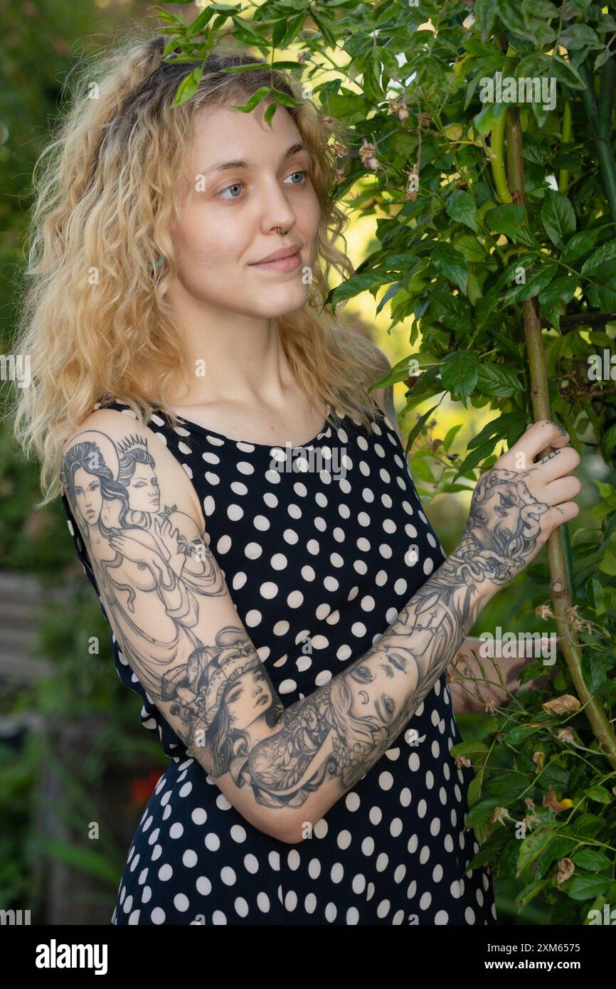 una donna bionda con tatuaggi che si aggrappano al ramo di un albero in un giardino estivo Foto Stock