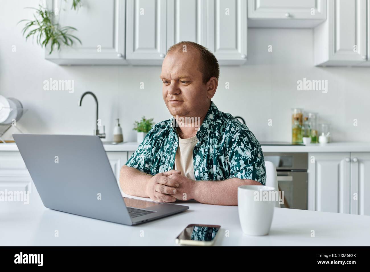 Un uomo con inclusività siede a un tavolo in una cucina moderna e lavora su un notebook. Foto Stock