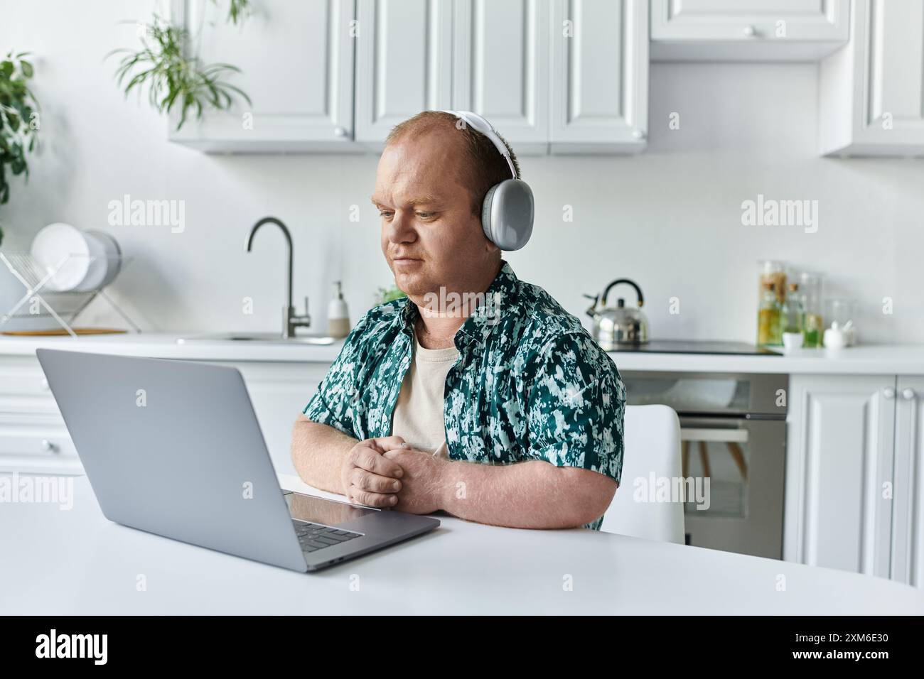 Un uomo con inclusività siede a un tavolo in cucina, indossa le cuffie e si concentra sul suo laptop. Foto Stock