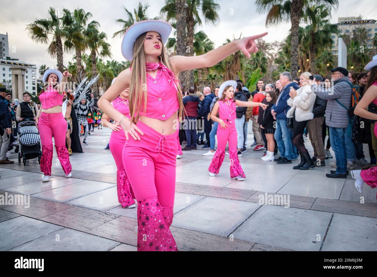 Una vivace esibizione di danza all'aperto con Barbie, ballerina principale in rosa e travolgente vestita da letto, mette in posa il pubblico raccolto tra le palme sotto una nuvolosa Foto Stock
