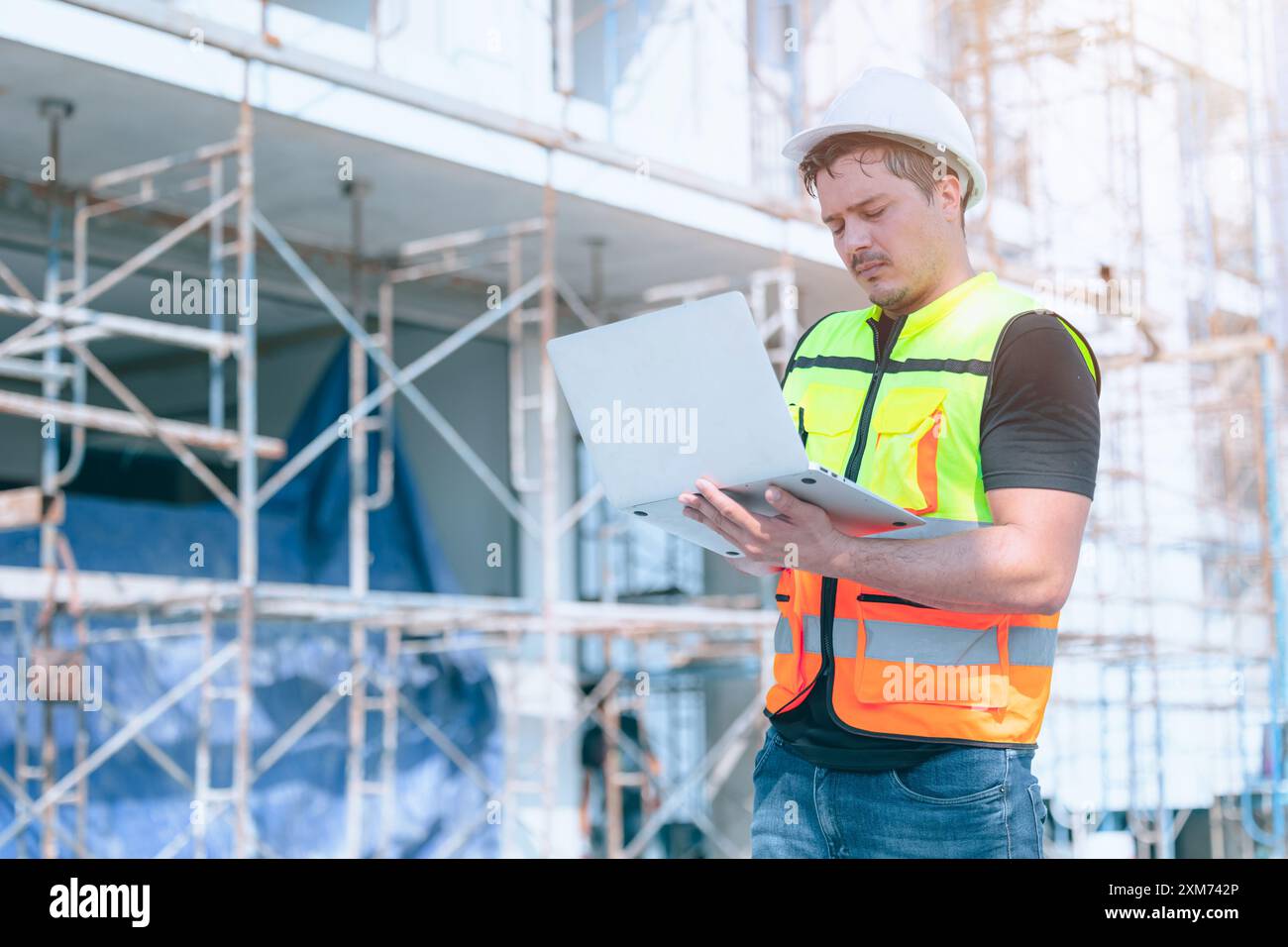 Operaio edile che indossa l'attrezzatura di sicurezza utilizzando un computer portatile in cantiere per gestire i dettagli del progetto. Foto Stock