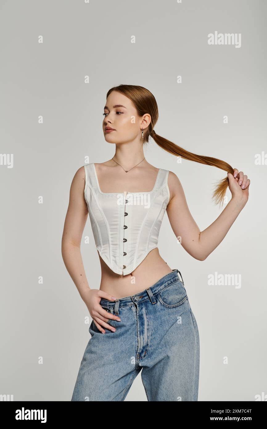 Una giovane donna posa con i capelli tirati indietro in una coda di cavallo, indossando un corsetto bianco e jeans blu. Foto Stock