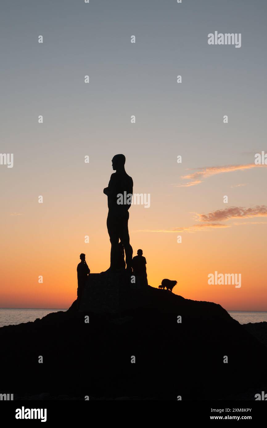 Grande statua di un uomo sulla riva dell'oceano Atlantico. La sua silhouette al tramonto, insieme a due persone e un cane. Cielo colorato, acqua calma. PA Foto Stock