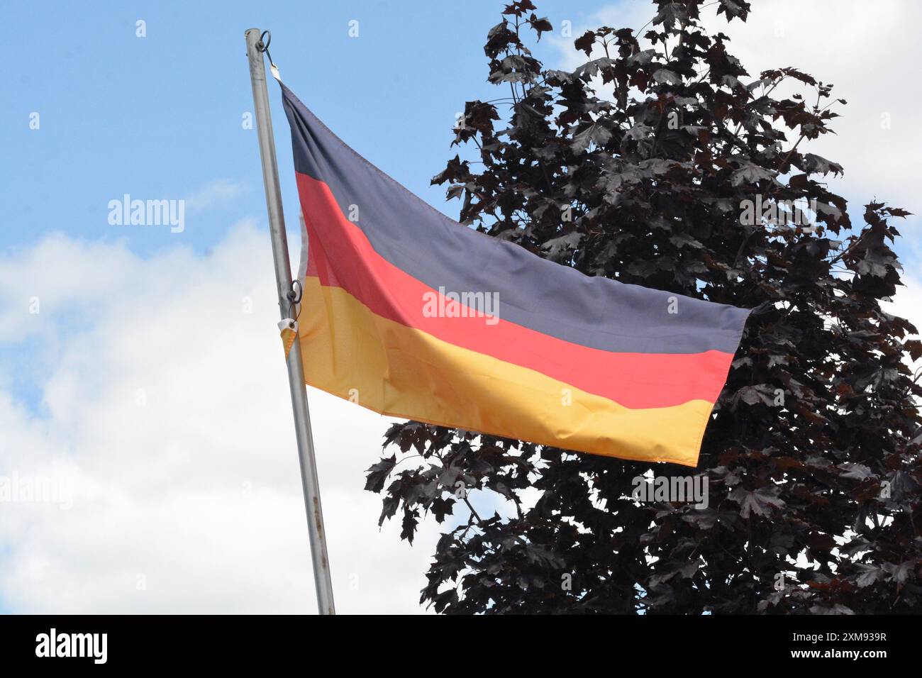 La bandiera nazionale della Germania vola su un palo in una splendida giornata estiva con uno sfondo di cielo blu e nuvole bianche. Foto Stock