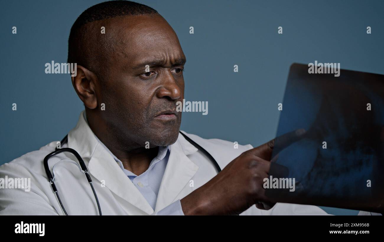 Medico maschio afroamericano pensivo maturo esaminare radiografia in sfondo blu studio uomo chirurgo traumatologo controllare risultati scansione analizzare lesioni su Foto Stock