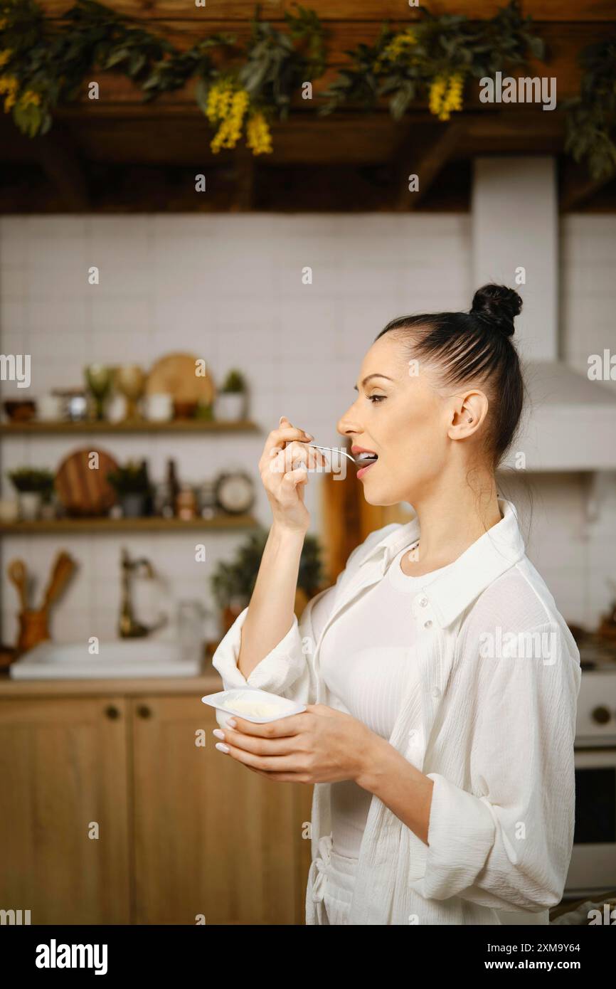 Donna con una camicia bianca in piedi di profilo in cucina a mangiare budino o yogurt Foto Stock