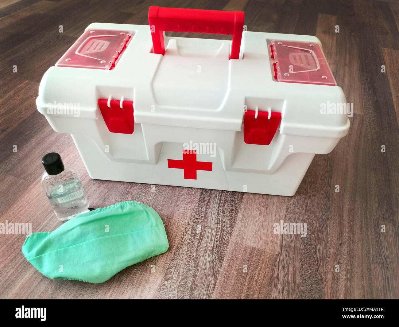 Scatola medica con una croce rossa, igienizzatore per le mani e maschera verde su un pavimento di legno, che rappresenta la preparazione e la sicurezza Foto Stock