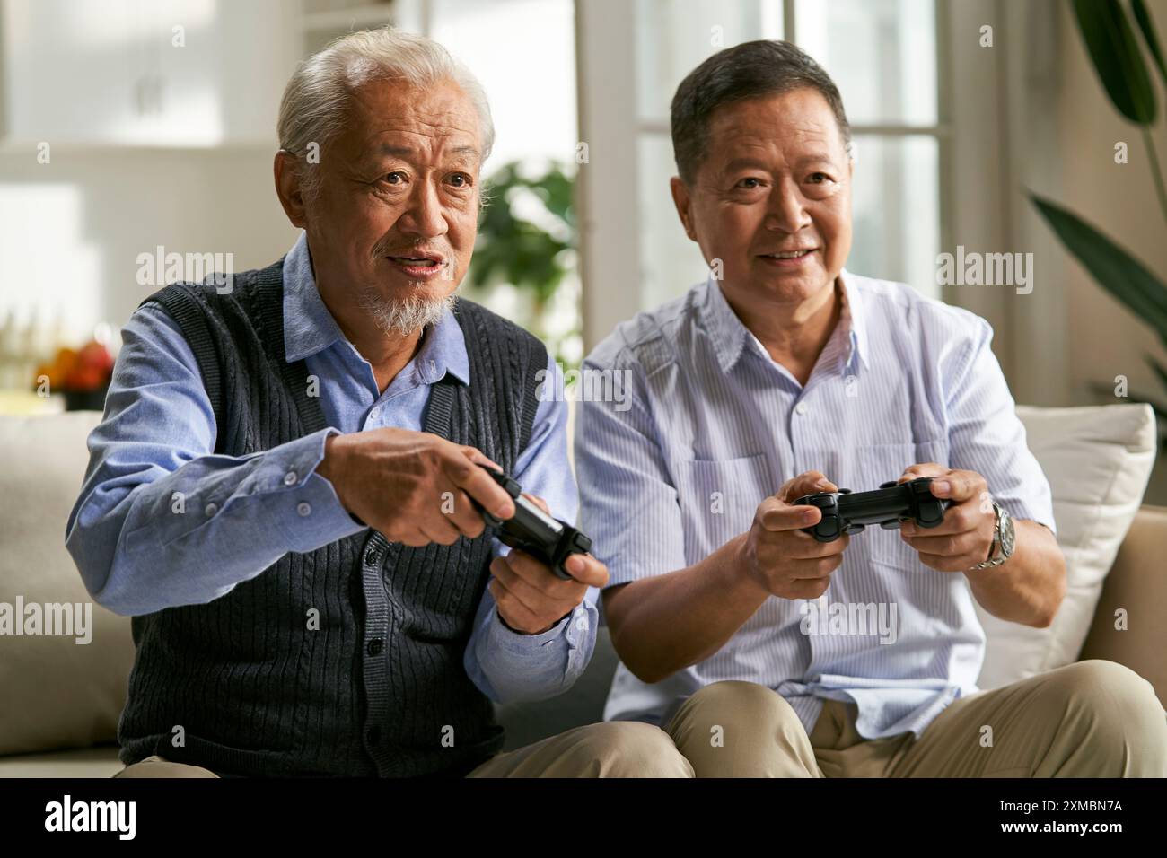 due uomini asiatici anziani felici seduti sul divano a casa a giocare a videogame Foto Stock