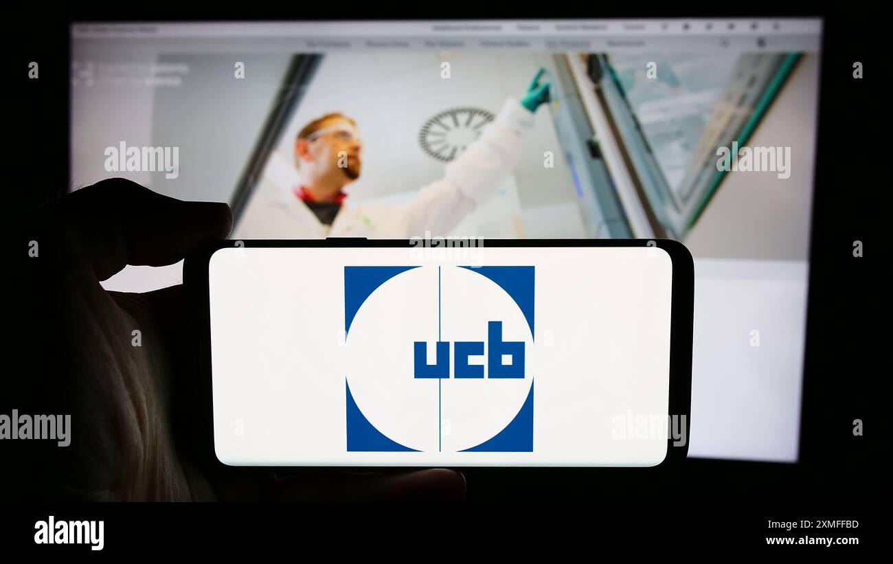 Persona che detiene un cellulare con il logo della società biofarmaceutica belga UCB S.A. di fronte alla pagina web aziendale. Mettere a fuoco il display del telefono. Foto Stock