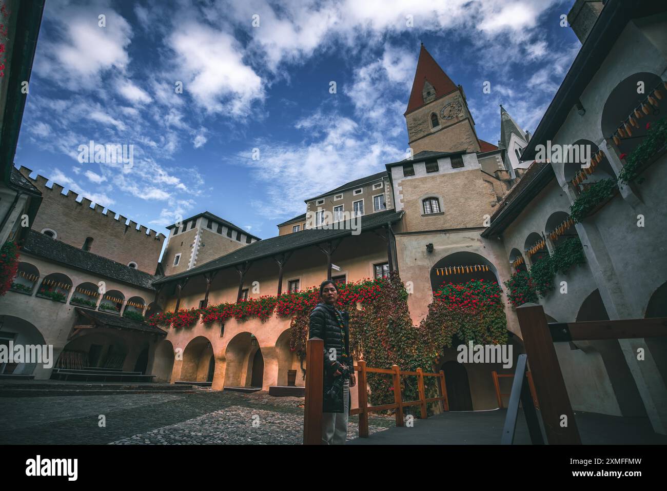 Austria - 26 ottobre 2023 : Una persona si trova nel cortile di un vecchio edificio con un passaggio ad arco e una torre sullo sfondo. C'è il rosso Foto Stock