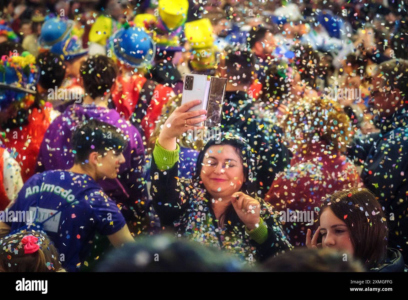 La folla celebrativa sfoggia cappelli colorati, abbraccia la festa e cattura momenti di gioia con gli smartphone in mezzo a docce di coriandoli. Foto Stock
