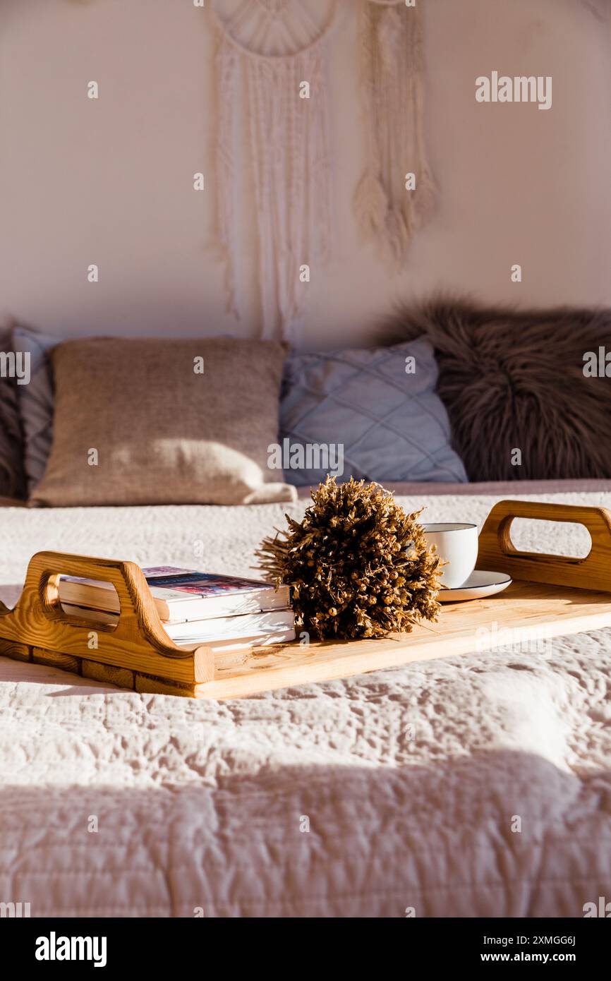 Accogliente camera da letto con letto illuminato dal sole, arredamento bohémien e vassoio in legno con libri e una tazza. Atmosfera invitante. Foto Stock