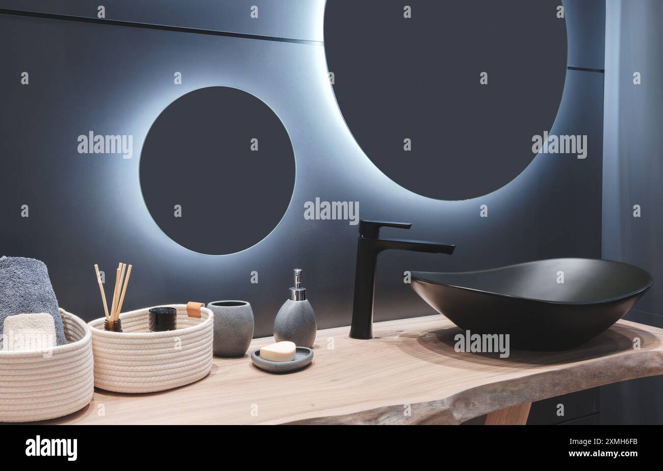 Interni lussuosi e moderni bagni blu con lavandino nero, specchio rotondo con illuminazione, lavandino nero con rubinetto, ripiano in legno naturale, circondato da s. Foto Stock