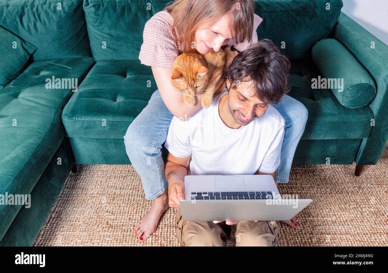 Coppia felice seduta su un divano con un laptop e sorridente, giovane coppia che fa una videochiamata, felice giovane famiglia con un gatto che fa una videoconferenza fr Foto Stock