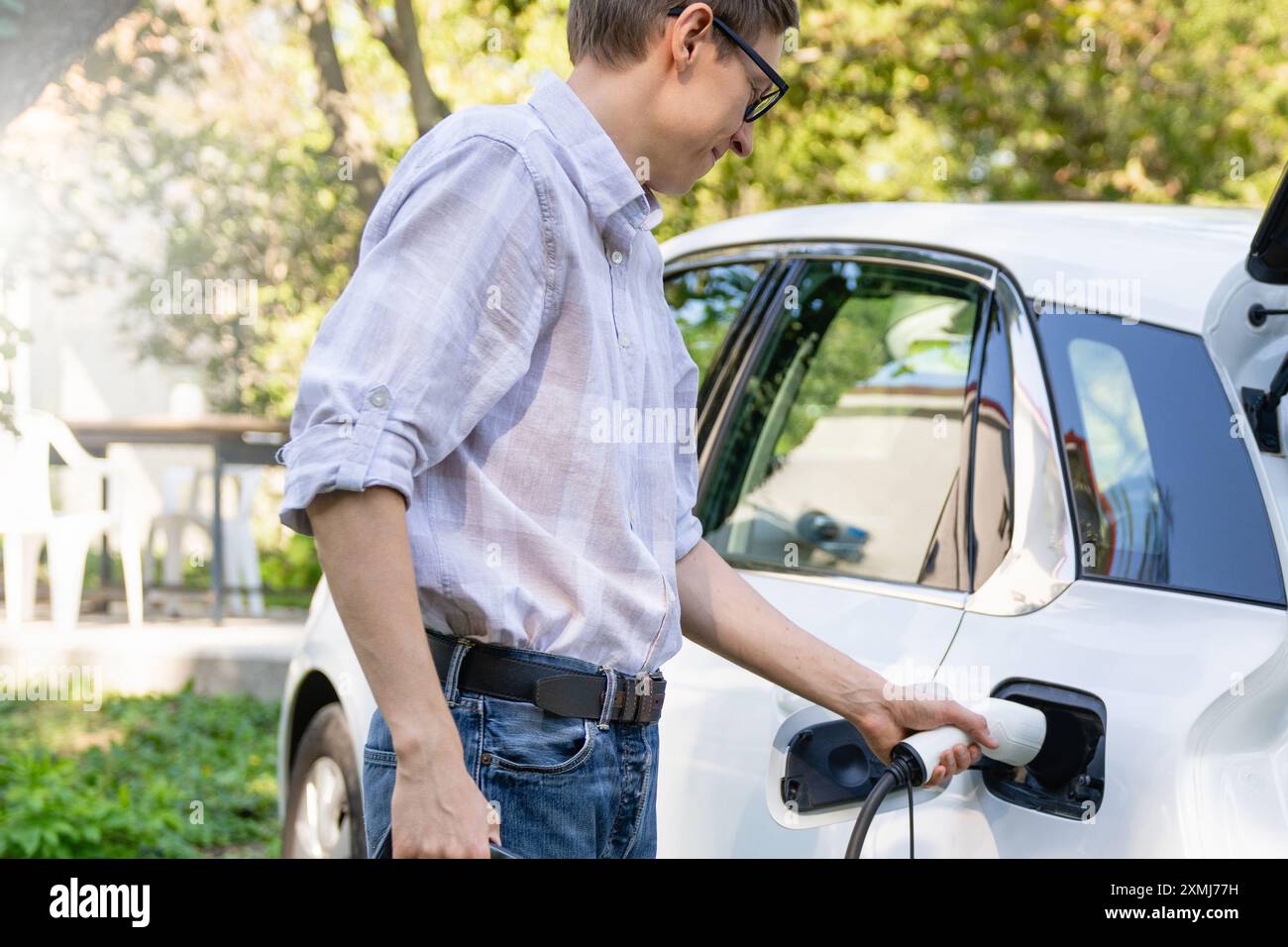 Uomo con una borsa per la spesa vicino a un'auto elettrica di ricarica nel cortile di una casa di campagna. Foto Stock