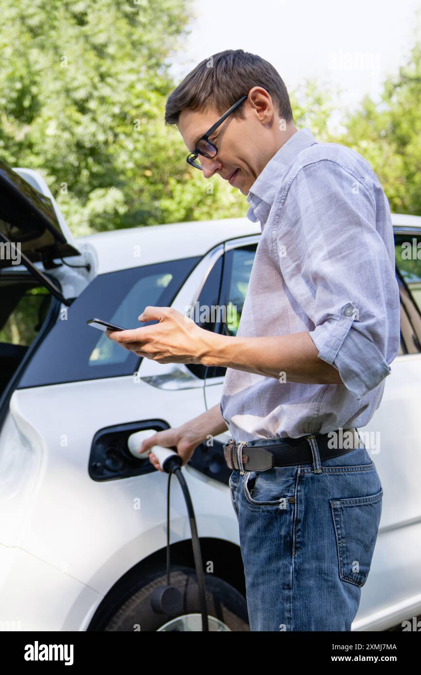 Uomo con una borsa per la spesa vicino a un'auto elettrica di ricarica nel cortile di una casa di campagna. Foto Stock