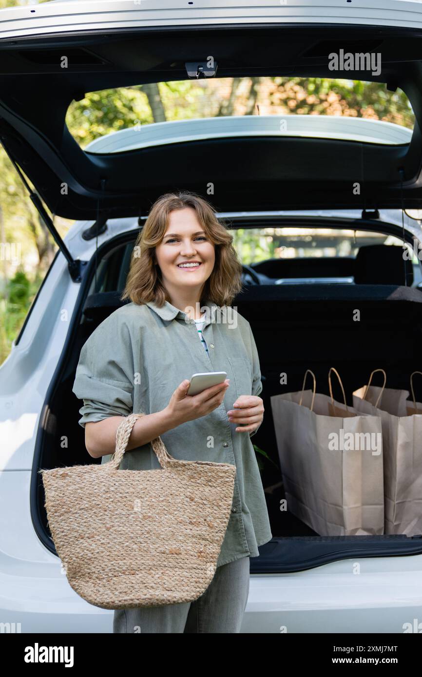 Donna con borsa della spesa accanto a un'auto elettrica in carica nel cortile di una casa di campagna. Foto Stock