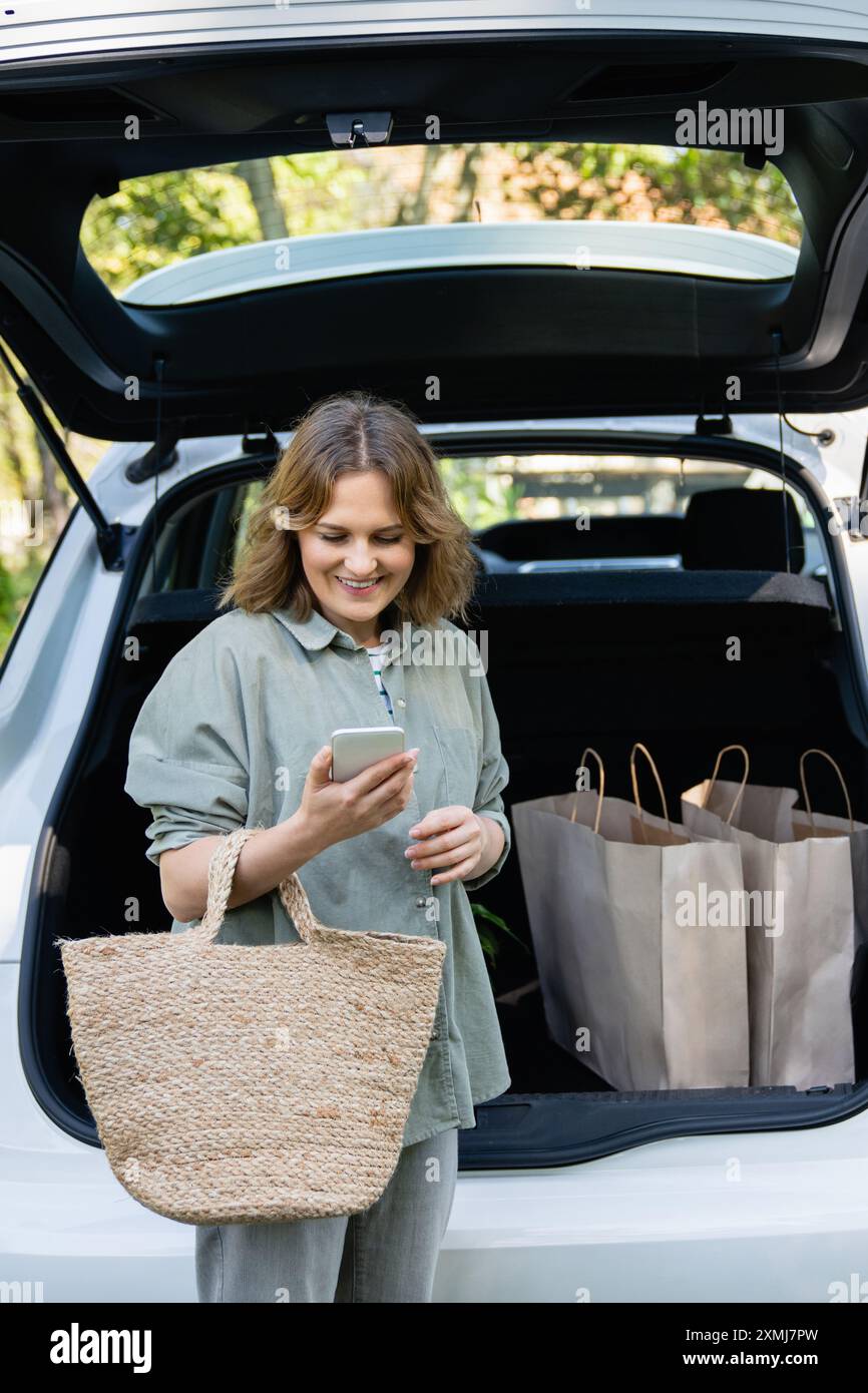 Donna con borsa della spesa accanto a un'auto elettrica in carica nel cortile di una casa di campagna. Foto Stock