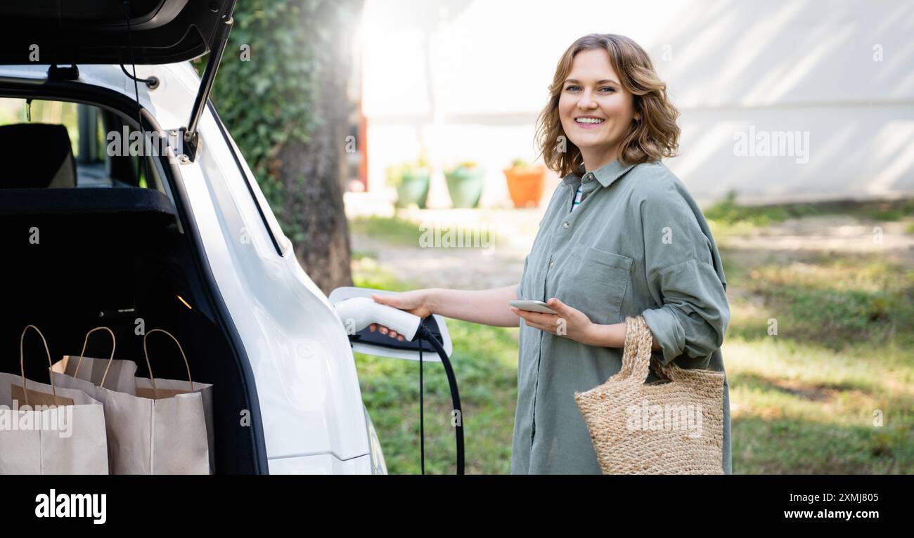 Donna con borsa per la spesa accanto a un'auto elettrica di ricarica nel cortile di una casa di campagna. Foto Stock