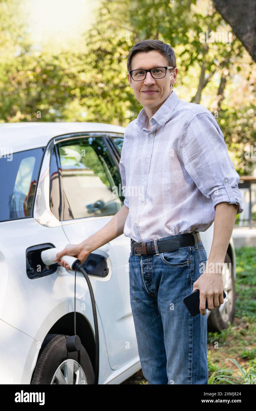 Uomo accanto a un'auto elettrica in carica nel cortile di una casa di campagna. Foto Stock