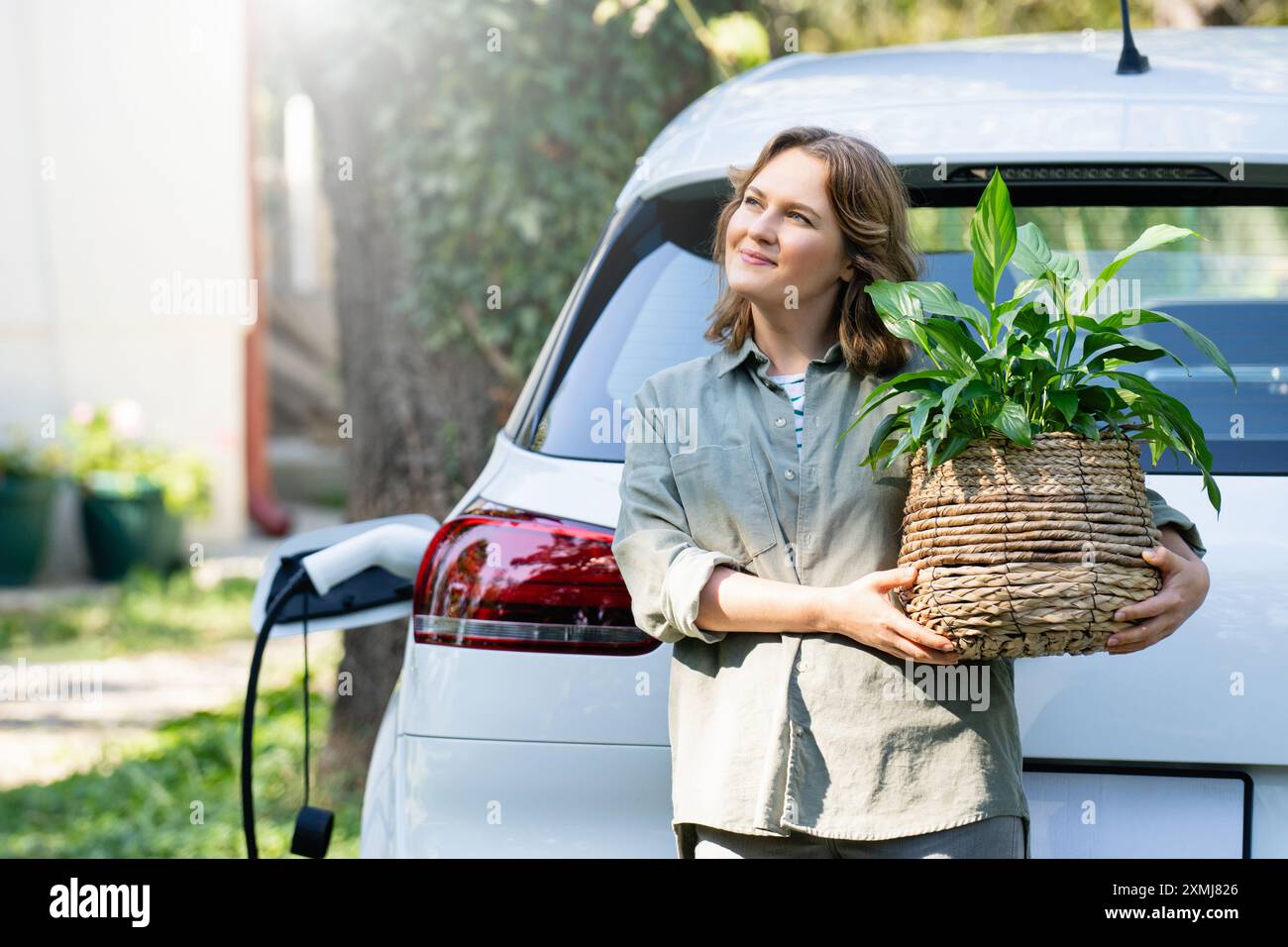 Donna con impianto in vaso vicino a un'auto elettrica di ricarica nel cortile di una casa di campagna. Foto Stock