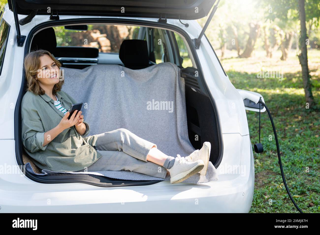 Donna con smartphone siede nel bagagliaio di un'auto elettrica. Foto Stock