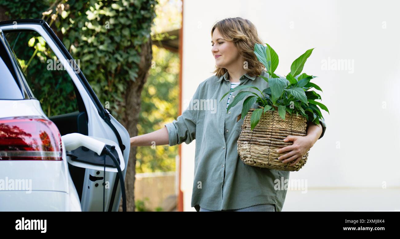 Donna con impianto in vaso vicino a un'auto elettrica di ricarica nel cortile di una casa di campagna. Foto Stock
