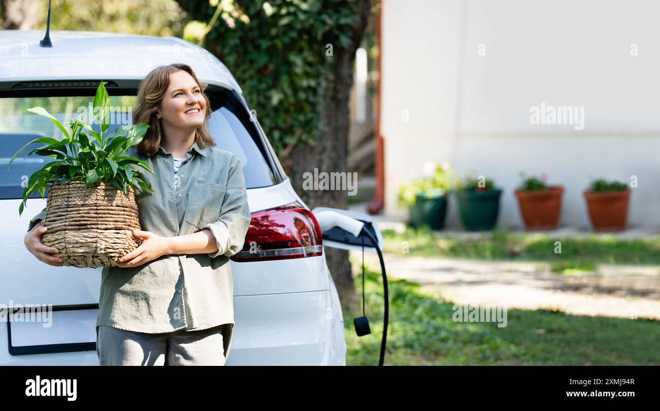 Donna con impianto in vaso vicino a un'auto elettrica di ricarica nel cortile di una casa di campagna. Foto Stock