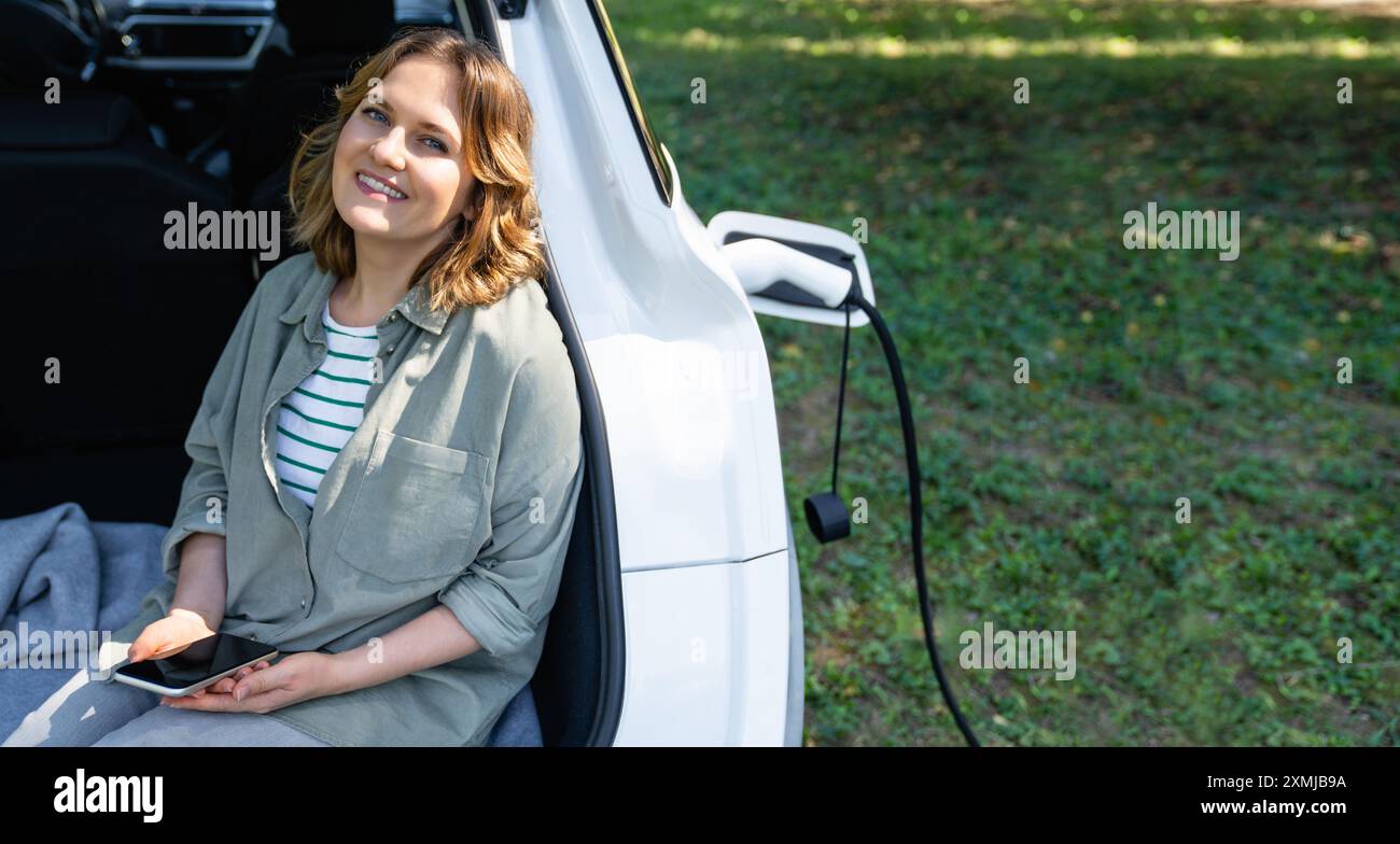 Donna con smartphone siede nel bagagliaio di un'auto elettrica. Foto Stock