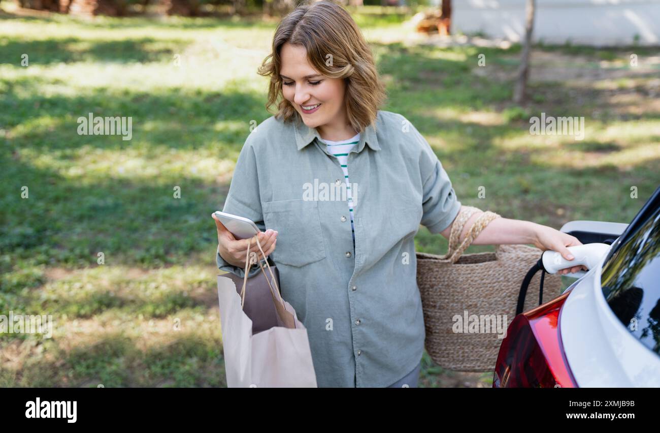 Donna con borsa della spesa accanto a un'auto elettrica in carica. Foto Stock