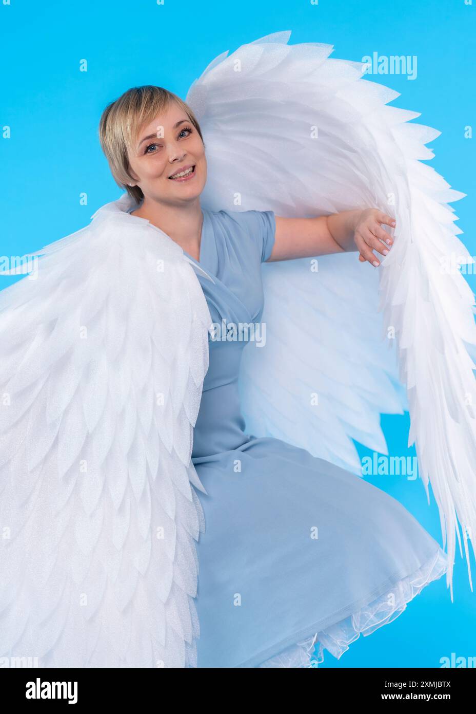 donna di mezza età in abito blu con grandi ali d'angelo seduta in studio su sfondo blu Foto Stock