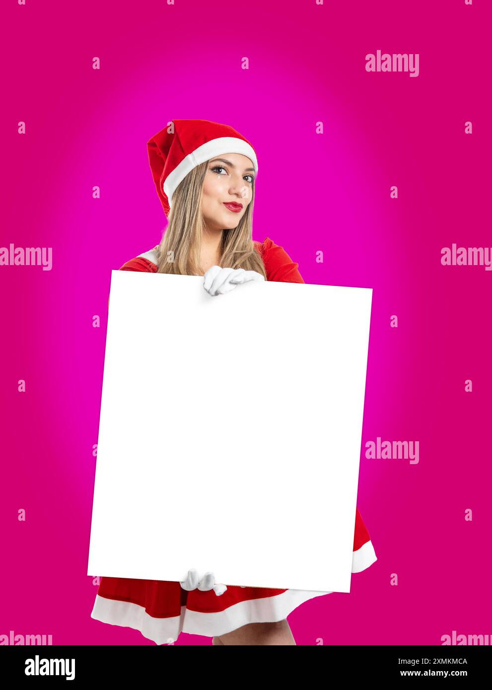 Bella giovane donna latina vestita da Babbo Natale che guarda la macchina fotografica e mostra un cartello come mock-up su sfondo magenta Foto Stock
